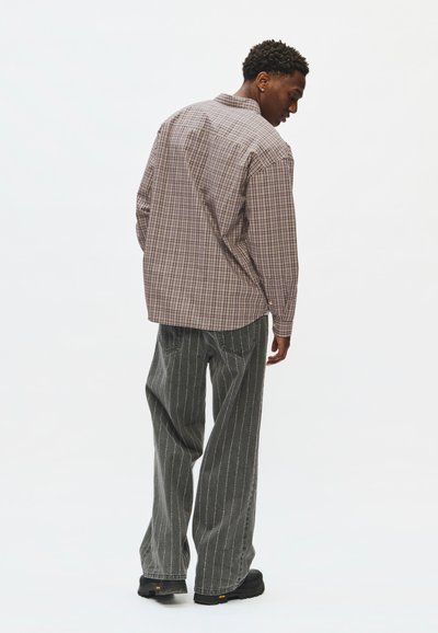 Homme portant une chemise à carreaux marron et un pantalon gris ample à fines rayures, debout et regardant vers le bas sur un fond blanc uni.