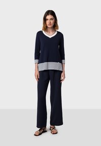 Maglietta a maniche lunghe blu navy con scollatura a V e orlo a righe, abbinata a pantaloni larghi blu navy. Il tessuto sembra morbido. Indossata con sandali neri.