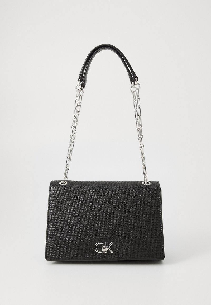 Sac à main noir texturé avec des bandoulières en chaîne argentée et un logo CK argenté sur le rabat avant, sur un fond clair uni.
