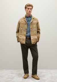 Giacca a fantasia tan con design geometrici, camicia blu a maniche lunghe, pantaloni verde oliva e scarpe mocassino beige. Texture lisce in tutto.