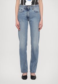 Šviesiai mėlyni tiesūs džinsai su nublukusiu finišu, penkių kišenių dizainu ir diržo kilpomis, dėvimi su grafine juoda ir balta marškinėliais.