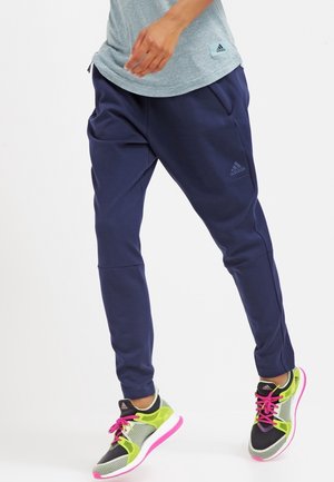 Personne portant un jogging Adidas bleu marine, un t-shirt Adidas gris, et des chaussures de course Adidas vert néon, noir et rose avec des lacets roses.