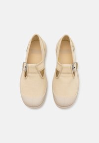 Zapatillas de lona beige claro con punta redonda, hebilla ajustable de tono plateado y suela de goma texturada. Presenta un diseño calado y un interior suave.