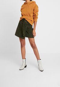 Mujer vestida con sudadera naranja, falda de pana verde oscura con cinturón, y botas blancas hasta el tobillo con patrón de serpiente, de pie con las piernas separadas.