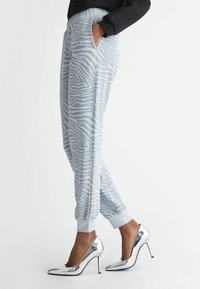Person med grå zebra-mönstrade joggers och silverfärgade högklackade skor, med en hand i fickan mot vit bakgrund.