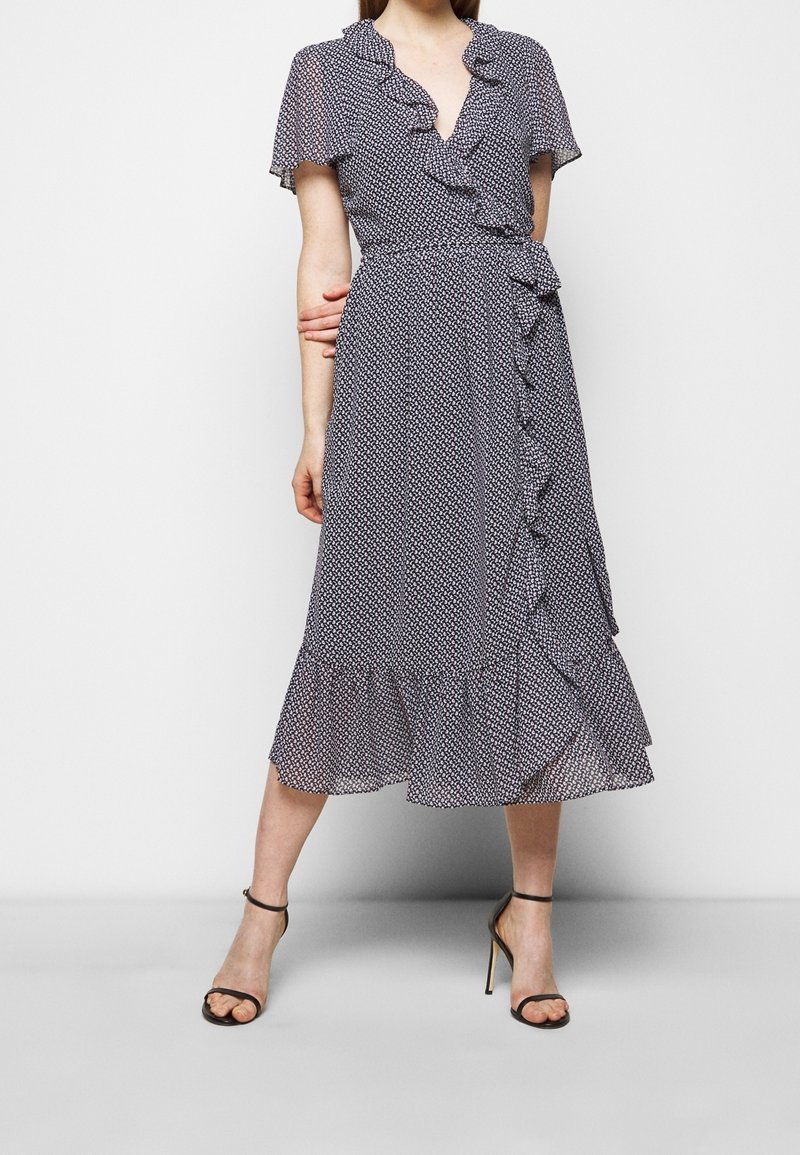 Femme portant une robe portefeuille midi bleu marine avec petits motifs blancs, détails volantés, manches courtes et sandales noires à talons hauts.