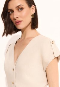 Beżowa sukienka z dekoltem w serek i krótkimi rękawami, z guzikami na ramionach. Modelka nosi złote kolczyki w kształcie promieni słońca. Gładka tekstura materiału.