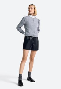 Žena stoji odjevena u sivi kardigan s gumbićima preko bijele bluze s visokim izrezom, crne kožne šortse, sive čarape i crne loferice.