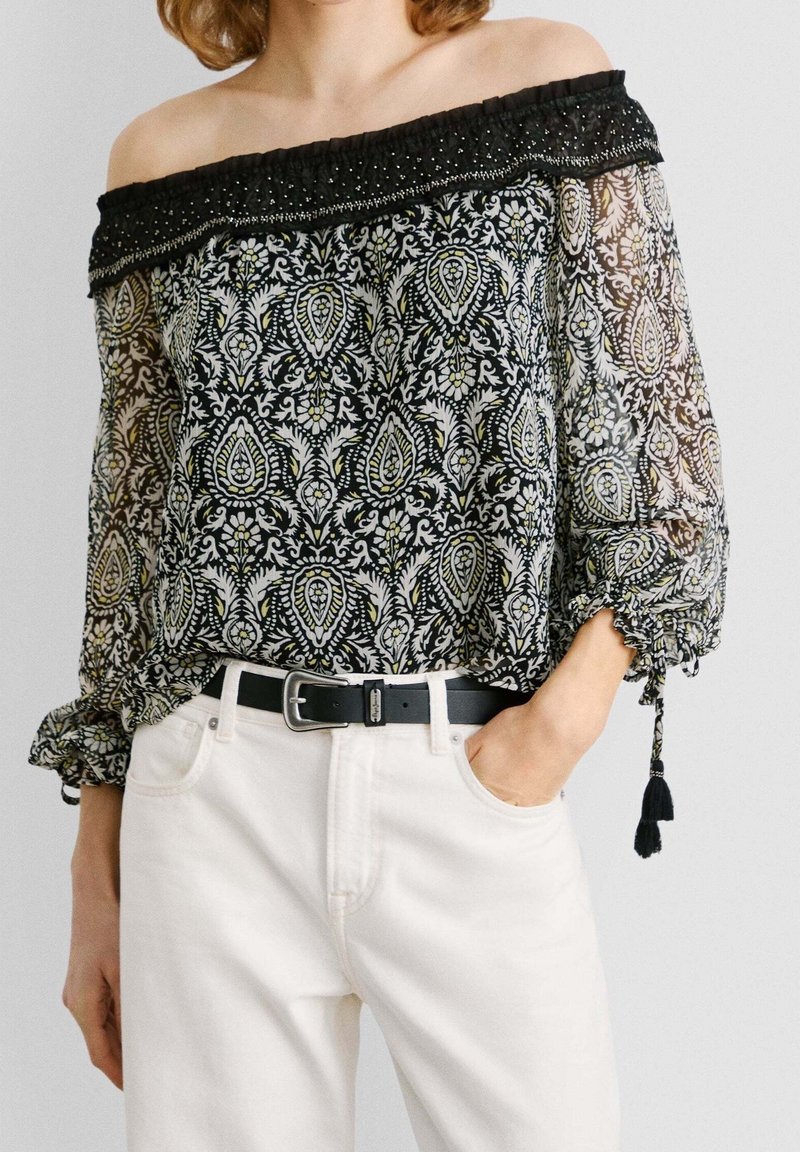 Femme portant un chemisier noir et blanc à motifs, épaules dénudées avec manches transparentes et un pantalon blanc avec une ceinture noire, une main dans la poche.