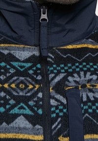 Pletená fleecová bunda s námořnickou horní částí a vzorovanou spodní částí v modrých, šedých a žlutých geometrických designech se zipem.