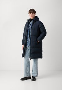 Navy puffer coat with a hood, featuring a zip front, two side pockets, and ribbed cuffs. Styled over a denim shirt and light jeans.