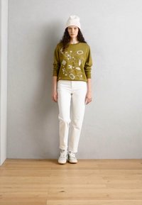 Pull olive avec broderie florale, jeans blancs et baskets de couleur claire. Le modèle se tient sur un sol en bois contre un mur gris.