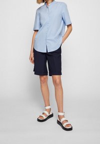 Hellblaues kurzärmeliges Hemd mit Knopfverschluss, kombiniert mit marineblauen, knielangen Shorts und weißen Plateausandalen mit mehreren Riemen.