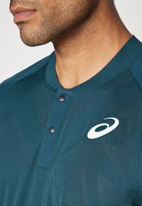 Teal sportshirt met een textuurdesign, korte kraag en knoopaccenten. Heeft een wit logo aan de linkerkant. De stof lijkt ademend te zijn.