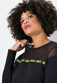 Zwarte lange mouw top met een mesh halslijn en groene tekstdetails. Zachte stof, relaxte pasvorm en textuuraccenten in mesh.
