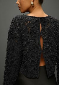 Zwart kant top met bloemenapplicaties, lange mouwen en een uitsnijding met sleutelgat aan de achterkant. Glad textuur met gelaagde bloemenpatronen.