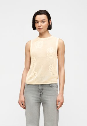 Γυναίκα με κοντά σκούρα μαλλιά, φοράει μπεζ αμάνικο τοπ από διχτυωτό ύφασμα με λουλουδάτα σχέδια και ανοιχτό γκρι τζιν, στέκεται μπροστά σε ένα απλό φόντο.