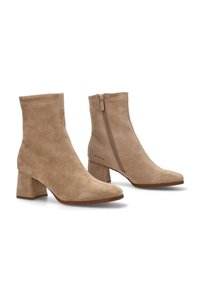 Botas de tobillo de ante beige con punta cuadrada y tacón cuadrado. Presentan un cierre lateral con cremallera y un logo en el lado exterior. Textura suave, diseño minimalista.