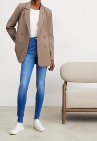 Blazer beige de doble botonadura, camiseta blanca, vaqueros skinny azules y zapatillas blancas. Tejido suave con detalles mínimos, en un fondo neutro.