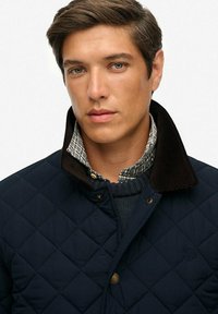 Chaqueta de punto azul marino con cuello de pana, llevada sobre una camisa de cuadros y un suéter de punto. Presenta detalles de botones y un logotipo.