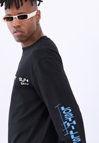Black long-sleeve t-shirt made of cotton, featuring white text on the chest and blue graphic text "LOST HILLS" on the left sleeve.