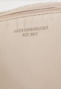 Beige fabric pouch with embossed text “ARKK COPENHAGEN EST. 2015” and subtle checkered texture, featuring a zipper along the top edge.