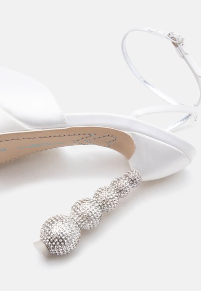 Λευκό πέδιλο με ψηλό τακούνι και λουράκι στον αστράγαλο, διακοσμημένο με πέντε μπαλάκια καλυμμένα με ασημένιες στρας σε φθίνουσα σειρά μεγέθους.