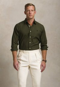 Roheline nööbitav särk terava kraega, koos kõrge talje ja volditud kreemjate pükstega. Aksessuaarideks on mustriga kaelaeht ja kell.