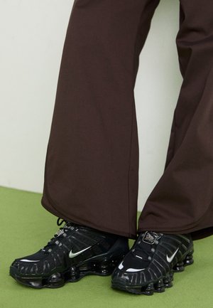 Osoba nosząca czarne buty Nike Shox z brązowymi spodniami stojąca na zielonej podłodze przed białą ścianą.
