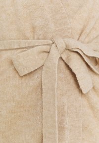 Beige stickad kavaj med en mjuk textur, som har ett knytbart tygskärp och en lös design. Stickmönstret är något synligt.