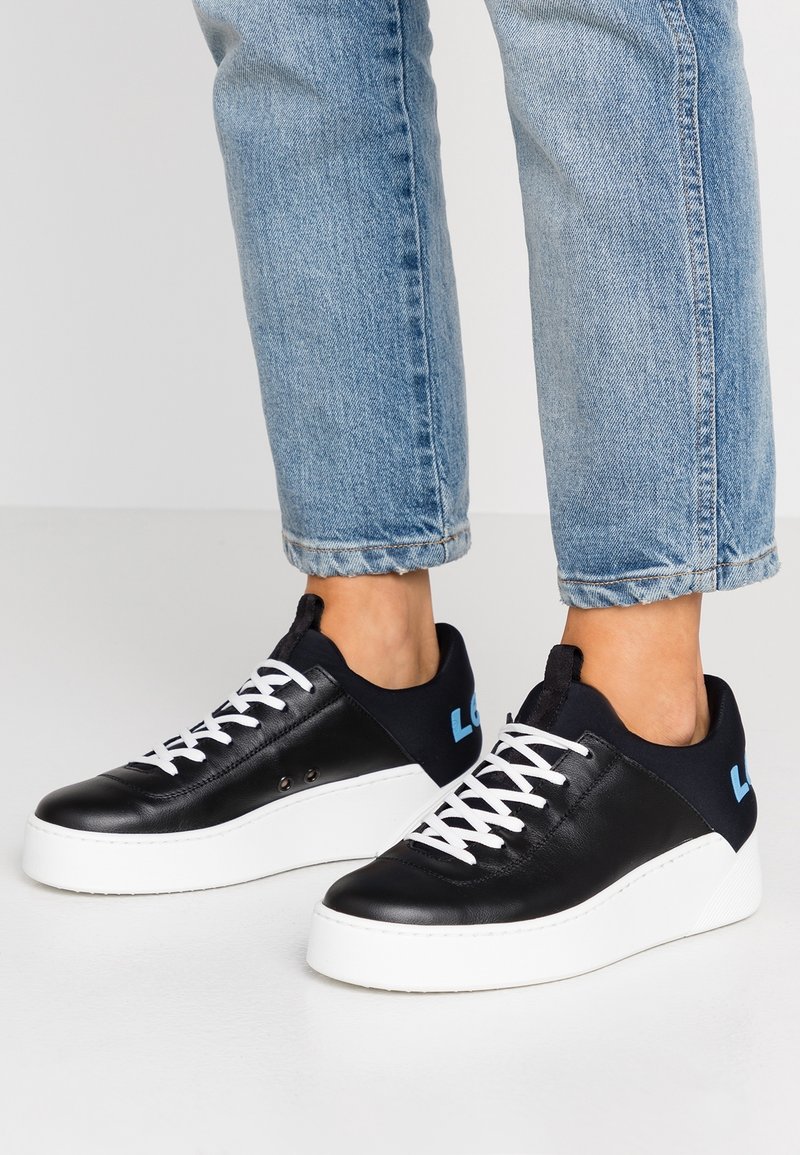 Personne portant un jean bleu clair et des baskets noires avec semelles et lacets blancs, debout sur une surface blanche.