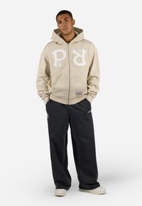 Beige zip-up hettegenser med hvit logo på brystet og frontlommer, parret med mørkegrå vide joggebukser og hvite sneakers.