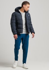 Marineblå puffjakke med en tætsiddende form, hætte og syede paneler, parret med en lys grå t-shirt og teal sweatpants. Hvide sneakers fuldender looket.