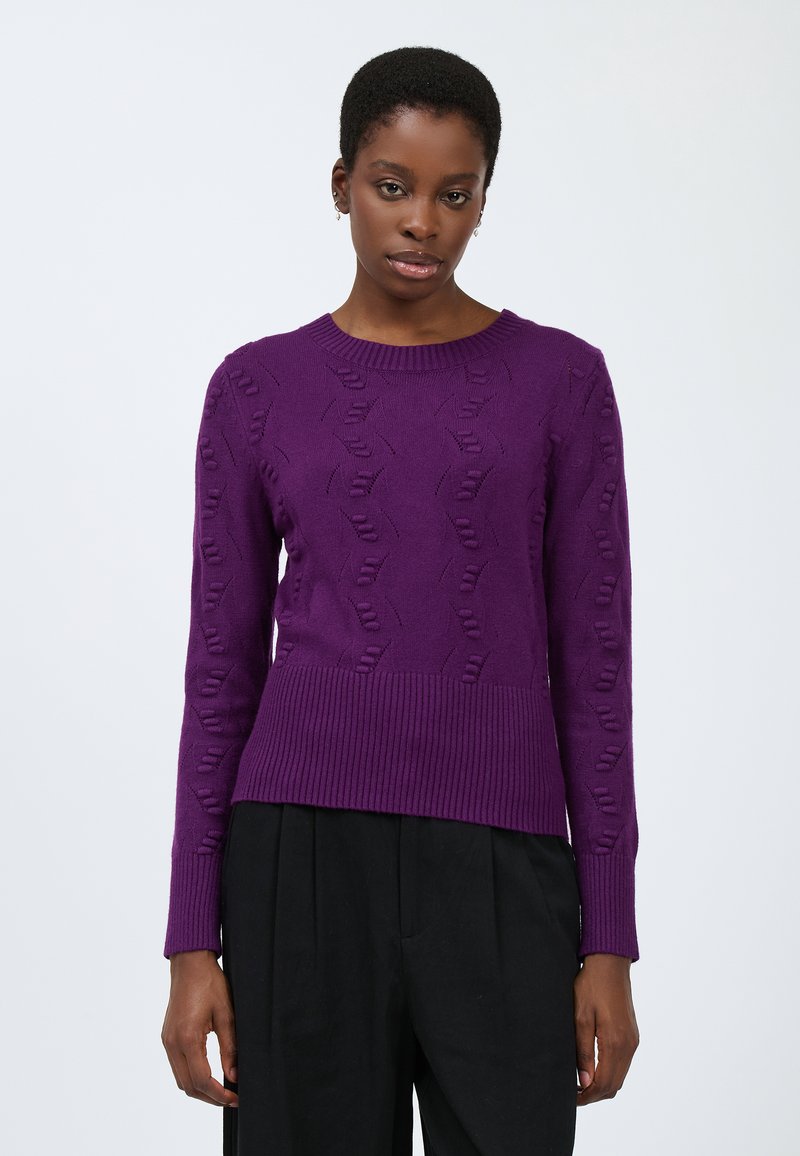 Lila gestrickter Pullover mit einem strukturierten Muster, rundem Halsauschnitt und geripptem Saum. Kombiniert mit schwarzen, plissierten Hosen.