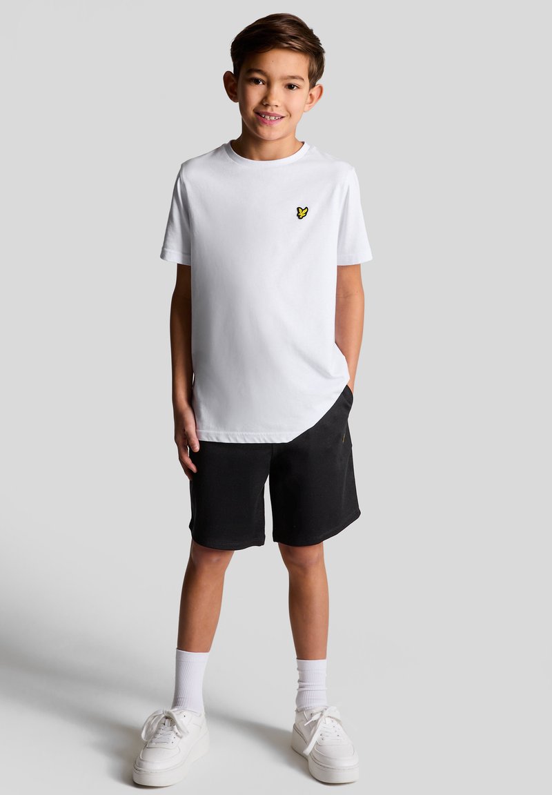 Hvid t-shirt med korte ærmer og et gult sommerfugl logo på brystet, parret med sorte shorts og hvide sneakers.