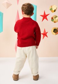 Pull rouge en tricot, pantalon en velours côtelé crème, bottines marron. En arrière-plan, des décorations géométriques aux couleurs pastel et métalliques.