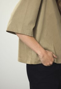 Kortærmet, beige bomuldsbluse med en afslappet pasform og synlige syninger, parret med sorte bukser, har en sidelomme i taljen.