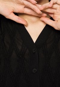 Černý cardigan s texturovaným diamantov�ým vzorem, má V-výstřih, dvě knoflíky a lehkou, průhlednou látku. Ruce jsou umístěny blízko výstřihu.
