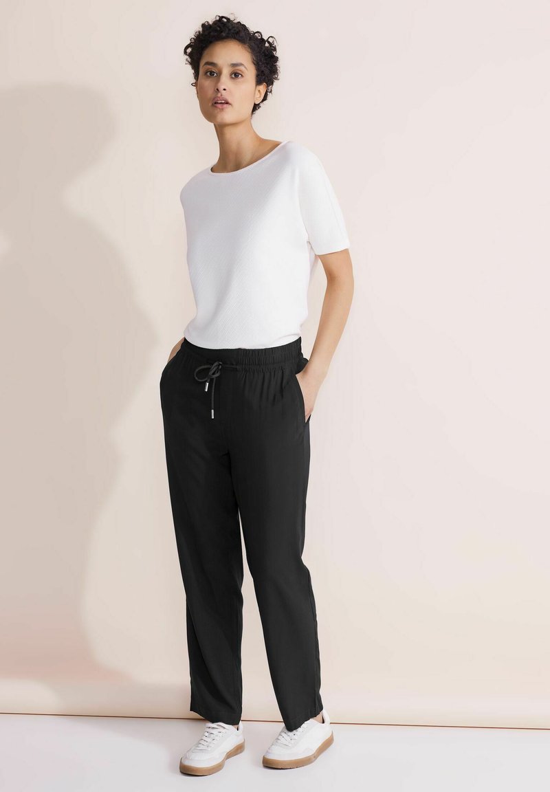 Vrouw staat tegen een beige muur en draagt een witte short-sleeve top, zwarte broek met trekkoord en witte sneakers met rubberen zolen.