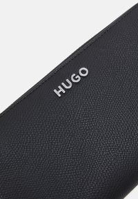 Svart läderpung med texturerad yta, med en silverfärgad "HUGO"-logotyp framtill. Rektangulär form, minimalistisk design.