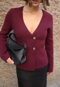 Bordowy sweter z dzianiny z głębokim dekoltem w kształcie litery V i dwoma złotymi guzikami. Połączony z czarną skórzaną torebką. Widoczna fakturowana tkanina.