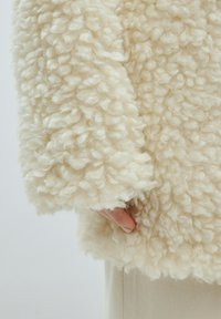 Crème-kleurige, puffy jas met een gestructureerde, krullende stof. Heeft losse mouwen en een zachte, gezellige uitstraling.