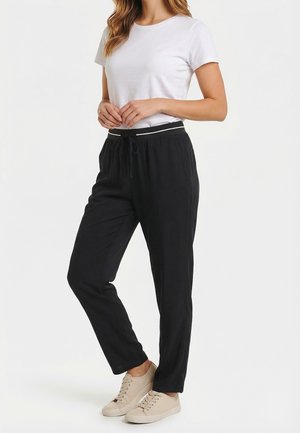 Donna che indossa una maglietta bianca a maniche corte, pantaloni neri con coulisse e striscia bianca in vita, e scarpe da ginnastica beige, in piedi davanti a uno sfondo bianco.