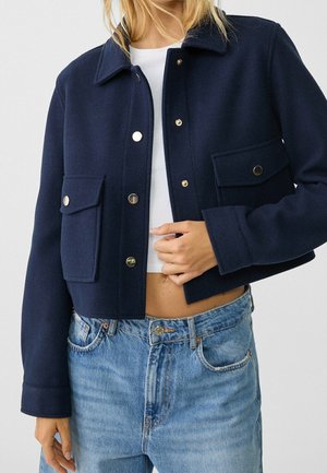 Nainen pukeutunut lyhennettyyn tummansiniseen napilliseen takkiin, jossa on läppätaskut, valkoisen lyhytpaita toppin ja vaaleansiniset farkut.