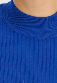 Pull bleu côtelé avec un col haut en maille. Le tissu est texturé, présentant des rayures verticales pour un aspect structuré.