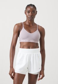 Mujer vistiendo un sujetador deportivo Nike lila y pantalones cortos deportivos Nike blancos, de pie contra un fondo claro liso.