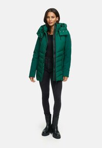 Groene pufferjack met een rits- en knoopsluiting, verstelbare capuchon en twee zijzakken, gedragen over zwarte slim-fit broek en zwarte laarzen.