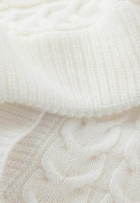 Biały dzianinowy materiał o prążkowanej fakturze i wzorach warkoczy, wykonany z miękkich, grubych włóczek, tworzący przytulny i ciepły wygląd.