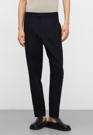 Mężczyzna w dopasowanych, czarnych, skróconych spodniach, beżowym prążkowanym topie i czarnych wsuwanych butach, stojący na jednolitym tle.