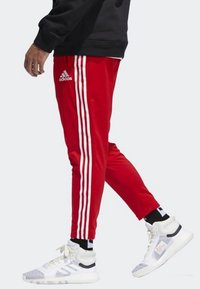 Rode sportbroeken met drie witte verticale strepen langs de zijkanten, gecombineerd met witte en grijze sneakers met zwarte accenten.